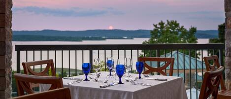 A table set for dining with blue glassware, positioned on a balcony overlooking a body of water and distant hills during sunset.