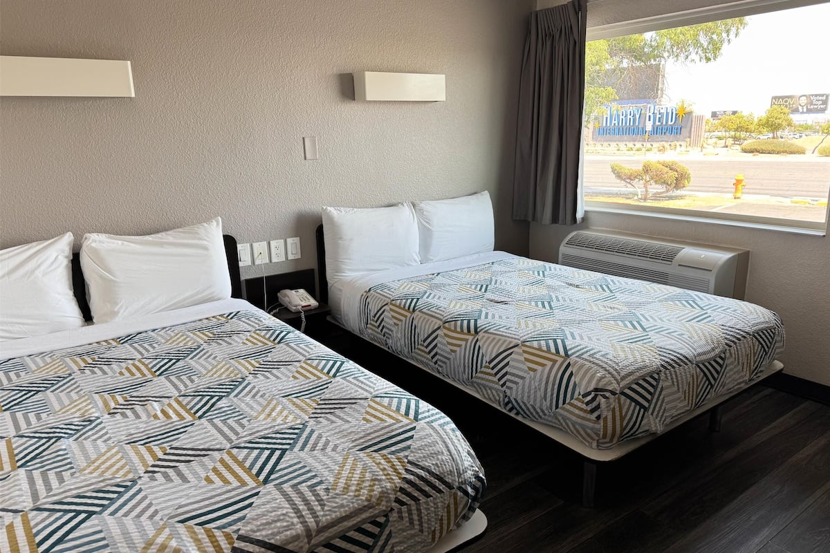 A hotel room with two beds, a window with a view of an airport, and a thermostat on the wall.