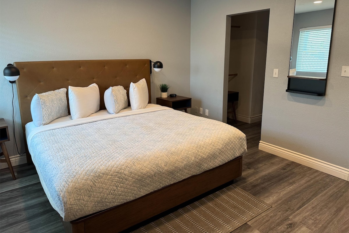 A bedroom with a large bed, a nightstand, a lamp, and a mirror on the wall.