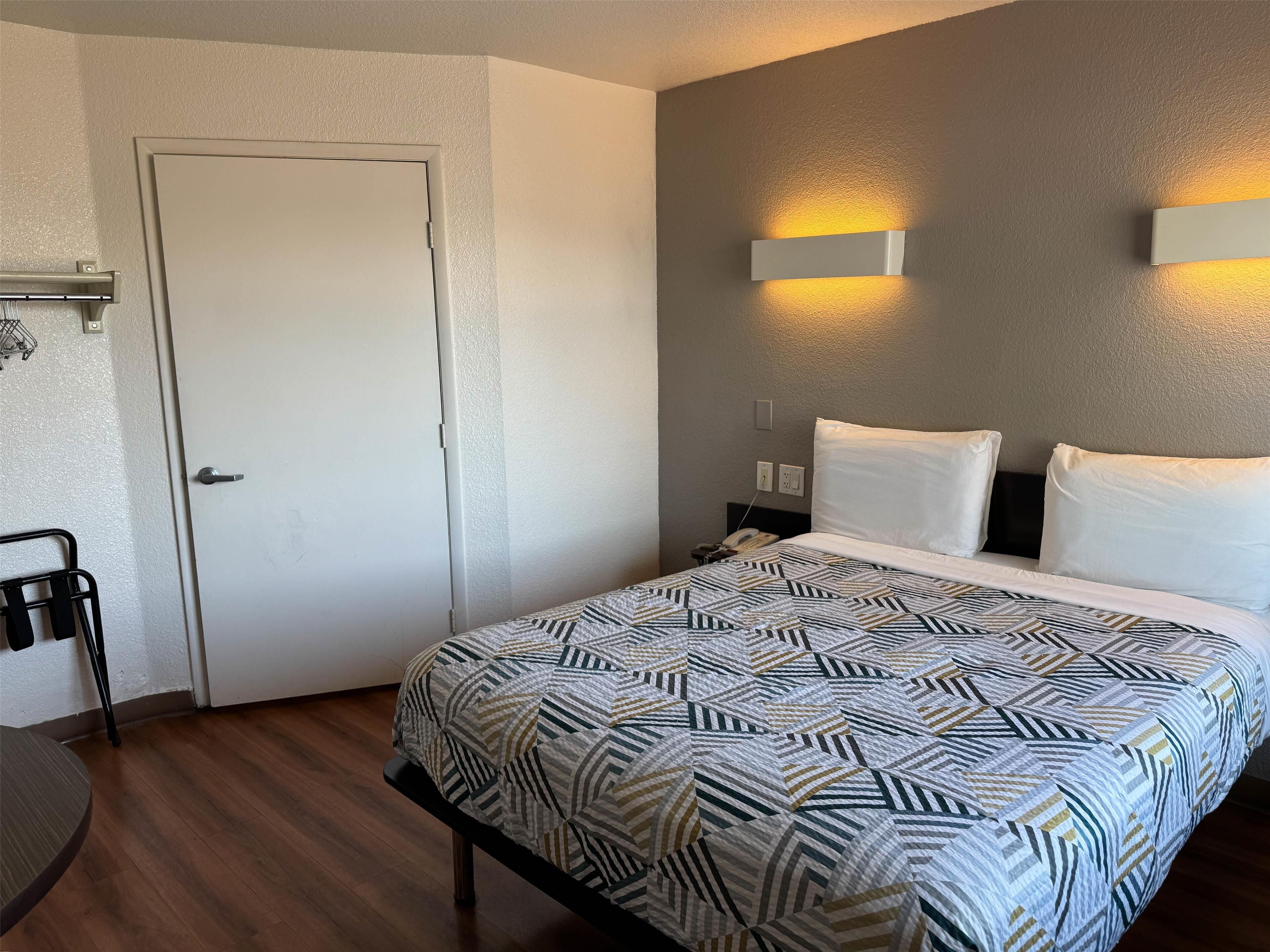 A hotel room with a bed, a desk, and a chair. There is a door, a wall-mounted light, and a phone on the nightstand.