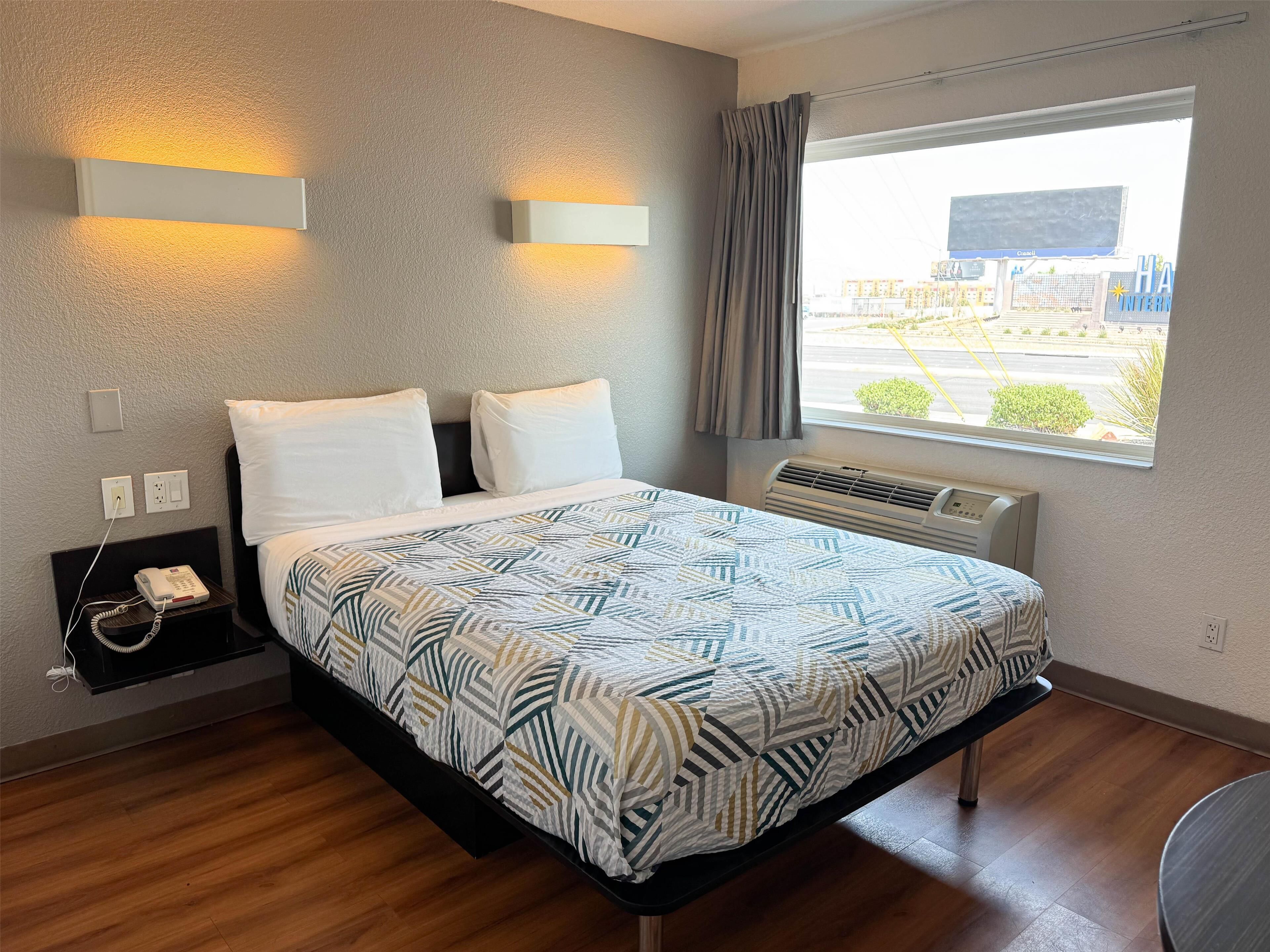 A hotel room with a bed, bedside table, phone, wall-mounted light, air conditioning unit, and a window with a view of the outside.