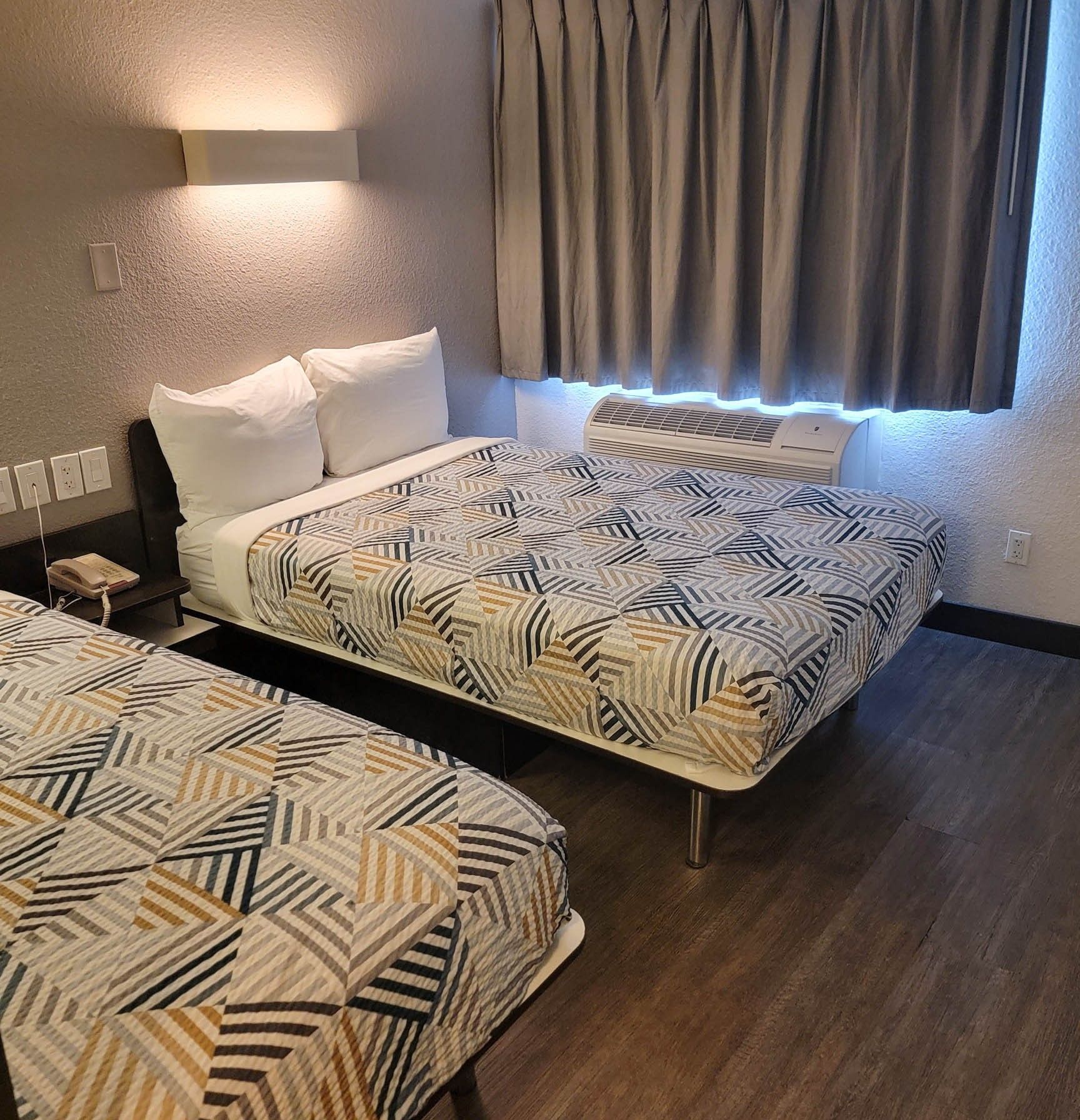 A hotel room with two beds, a bedside table with a phone, and a window with curtains.