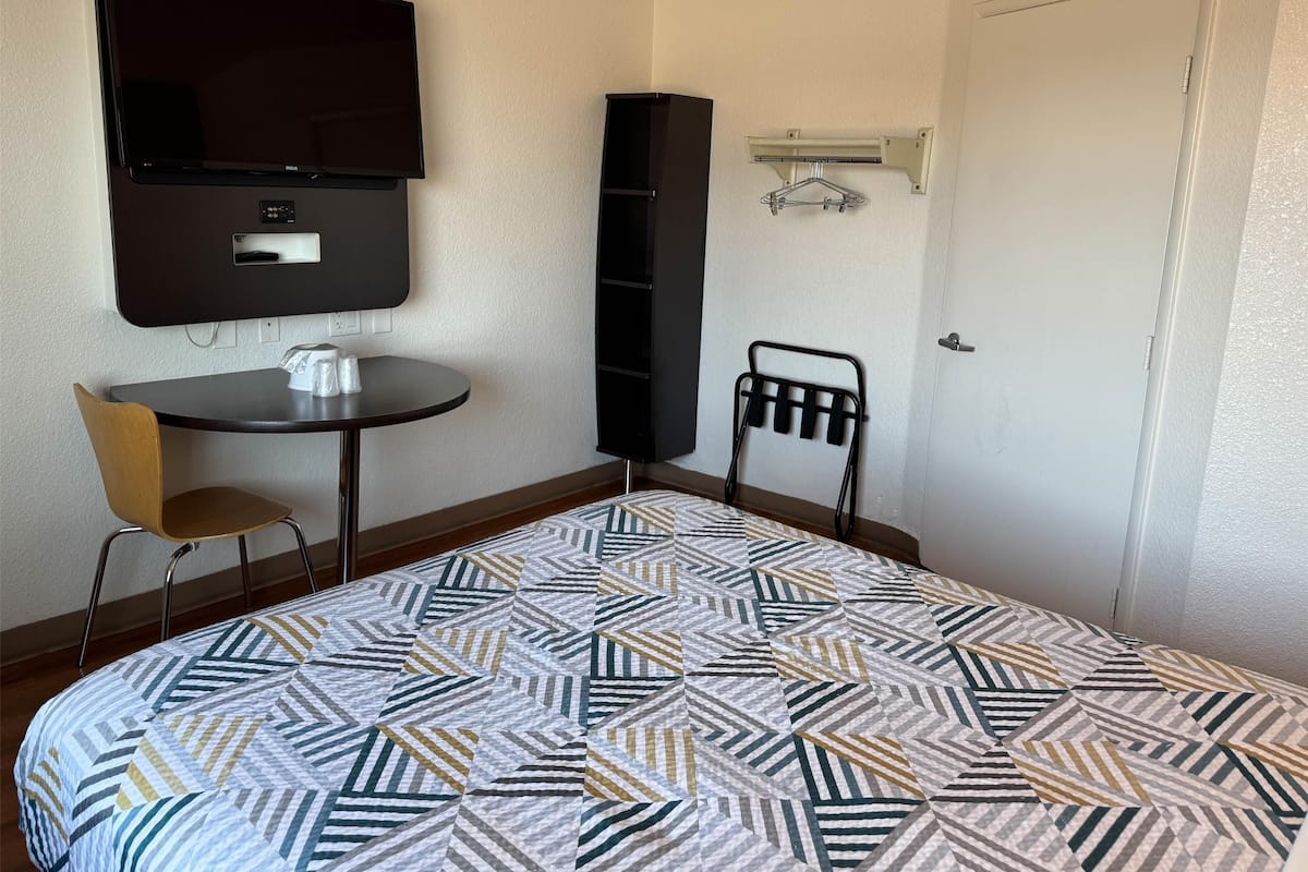 A hotel room with a bed, a TV mounted on the wall, a small table with a chair, and a coat rack.