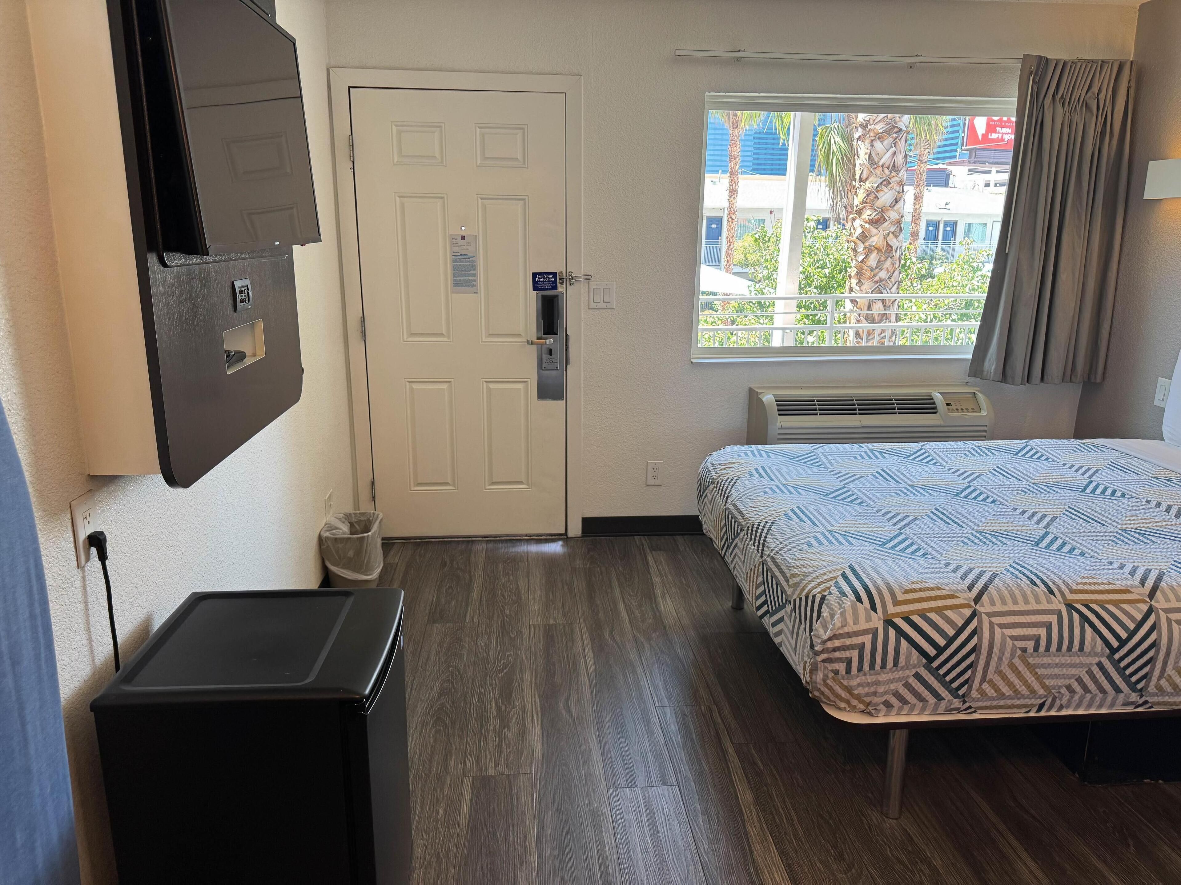 A hotel room with a bed, a desk, a TV, a window with curtains, and a door.