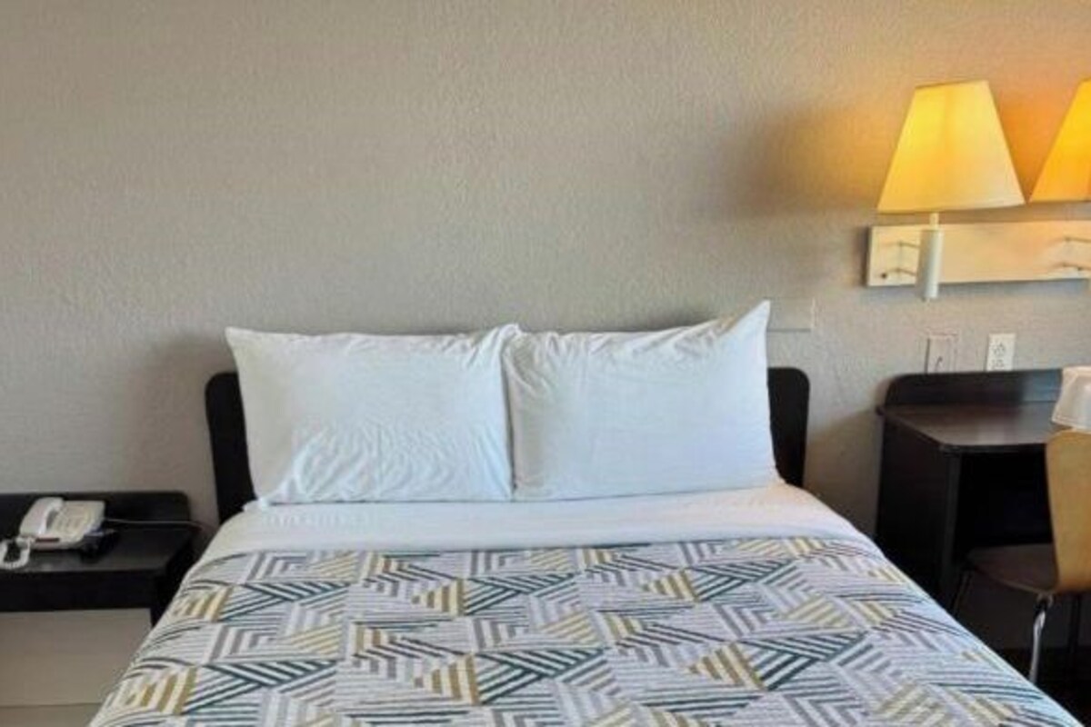 A hotel room with a bed, a nightstand with a phone, a desk with a lamp, and a chair.