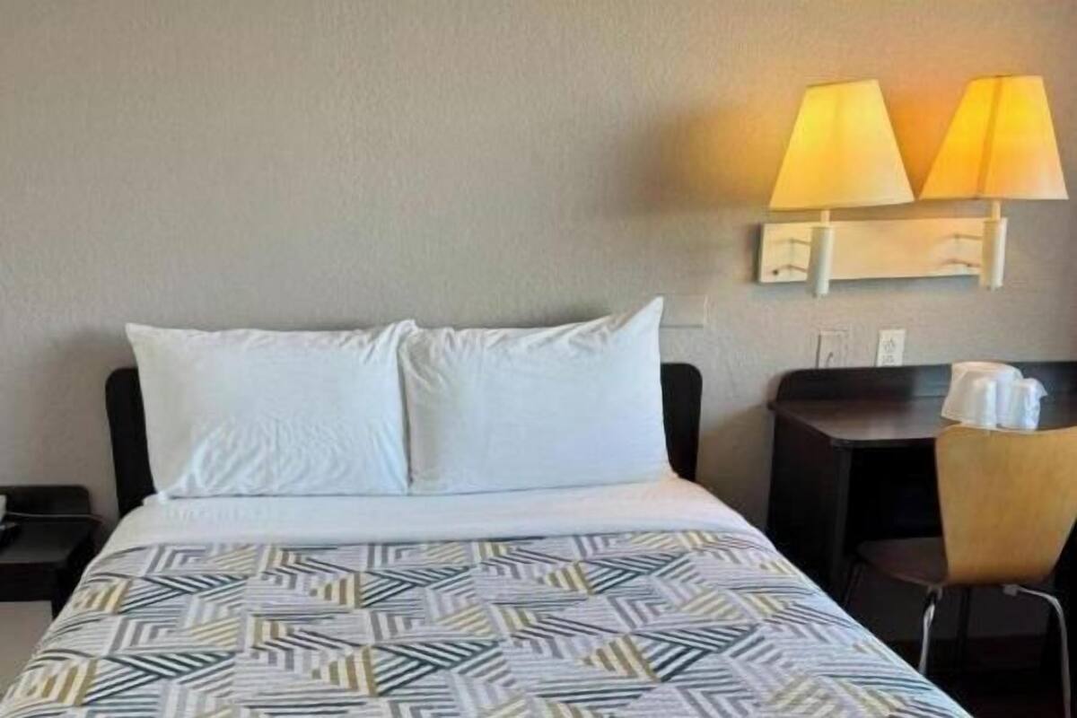 A hotel room with a bed, a nightstand with a phone, a desk with a lamp, and a chair.