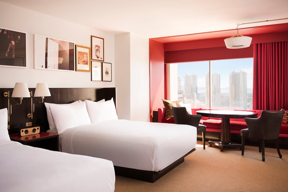 A hotel room with two beds, a dining table, and a sofa. There are framed pictures on the wall and a city view through the window.