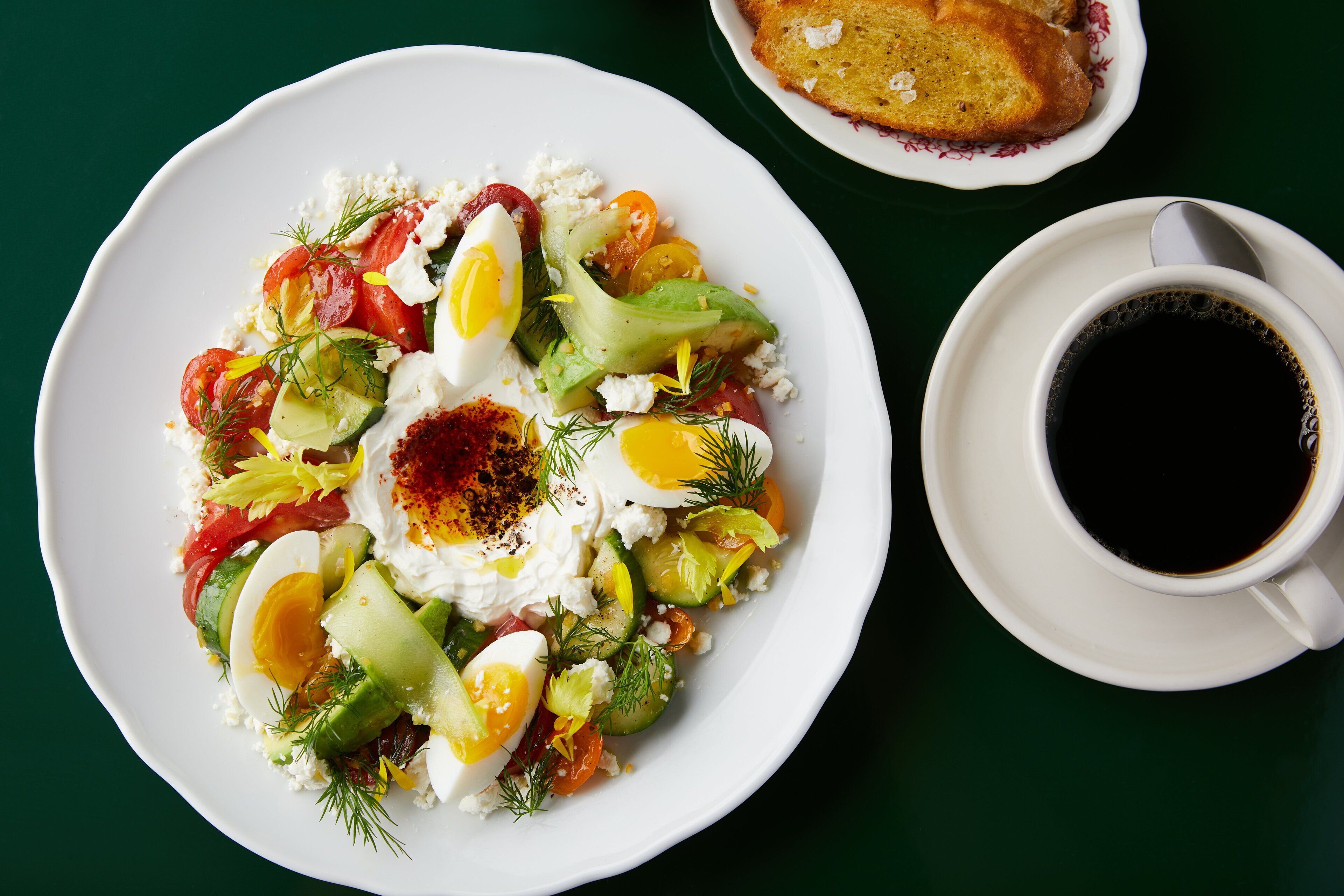 Une assiette de salade composée de laitue, d’œufs, de tomates, de concombres et de fromage, accompagnée d’une tasse de café et d’une tranche de pain, le tout posé sur une table verte.