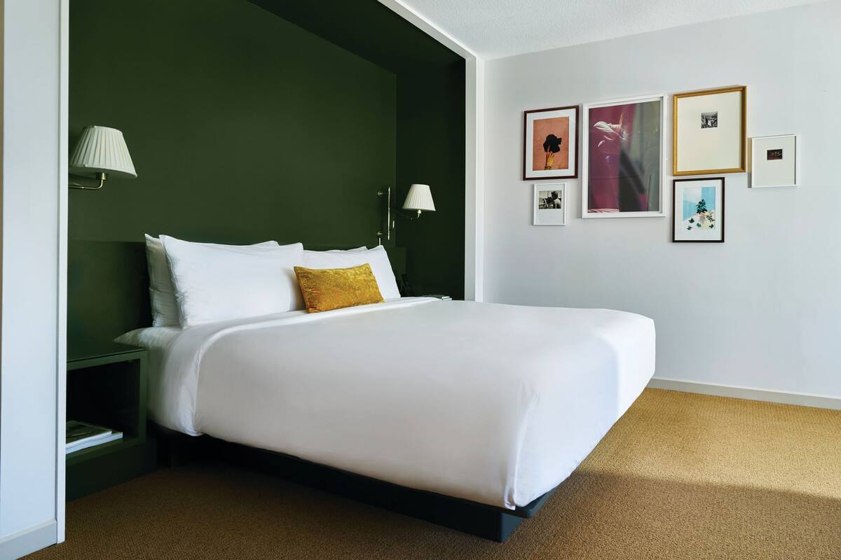 A neatly made bed with white linens and a single gold accent pillow, a nightstand with a lamp, and a wall with several framed pictures.