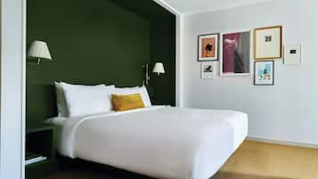 A neatly made bed with white linens and a single gold accent pillow, a nightstand with a lamp, and a wall with several framed pictures.
