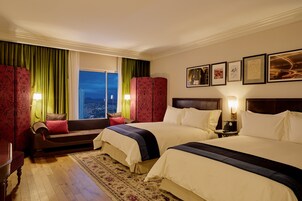 Camera d'albergo con due letti, un divano, un'ampia finestra con tende, vista notturna e quadri appesi alle pareti.