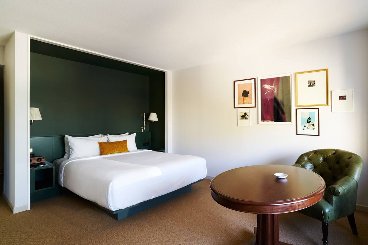 A hotel room with a bed, a round wooden table, a green armchair, and framed pictures on the wall.