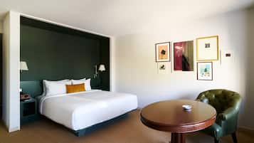 Una camera d'albergo con un letto, un tavolo rotondo in legno, una poltrona verde e quadri appesi alle pareti.