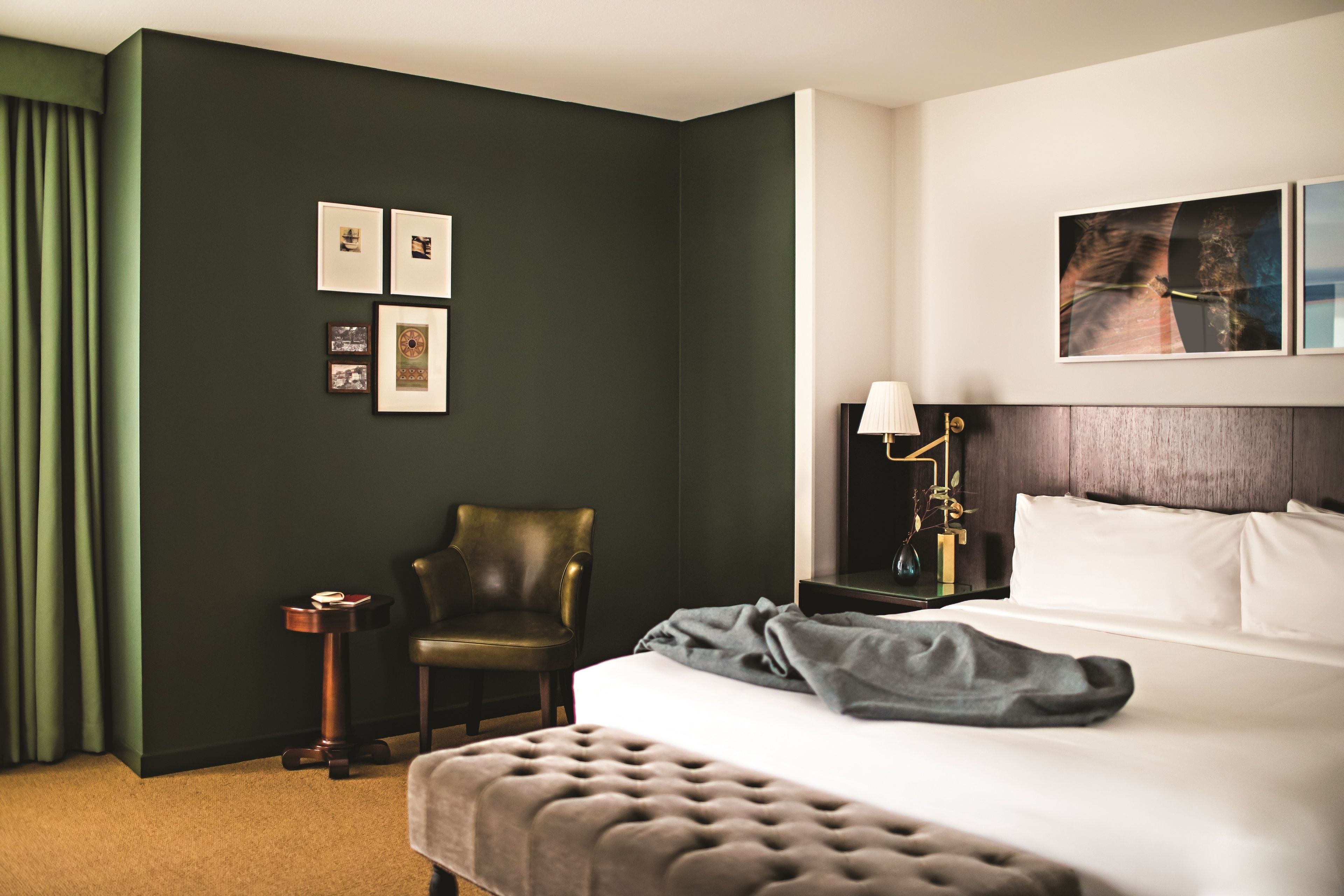 Habitación de hotel con una pared verde oscuro, una cama con ropa de cama blanca, un cabecero capitonado, una mesita de noche con una lámpara y cuadros enmarcados en la pared.