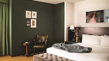 Habitación de hotel con una pared verde oscuro, una cama con ropa de cama blanca, un cabecero capitonado, una mesita de noche con una lámpara y cuadros enmarcados en la pared.