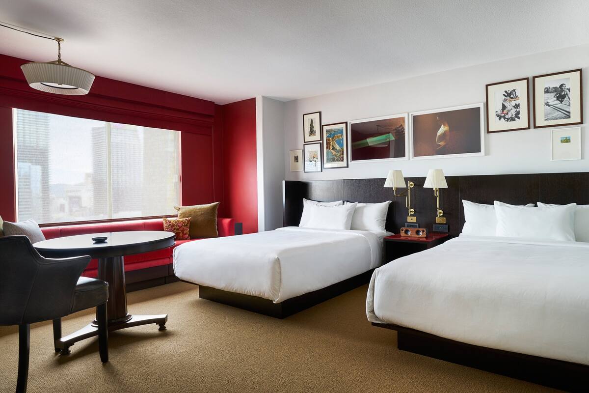 A hotel room with two beds, a desk, and a window with a city view.