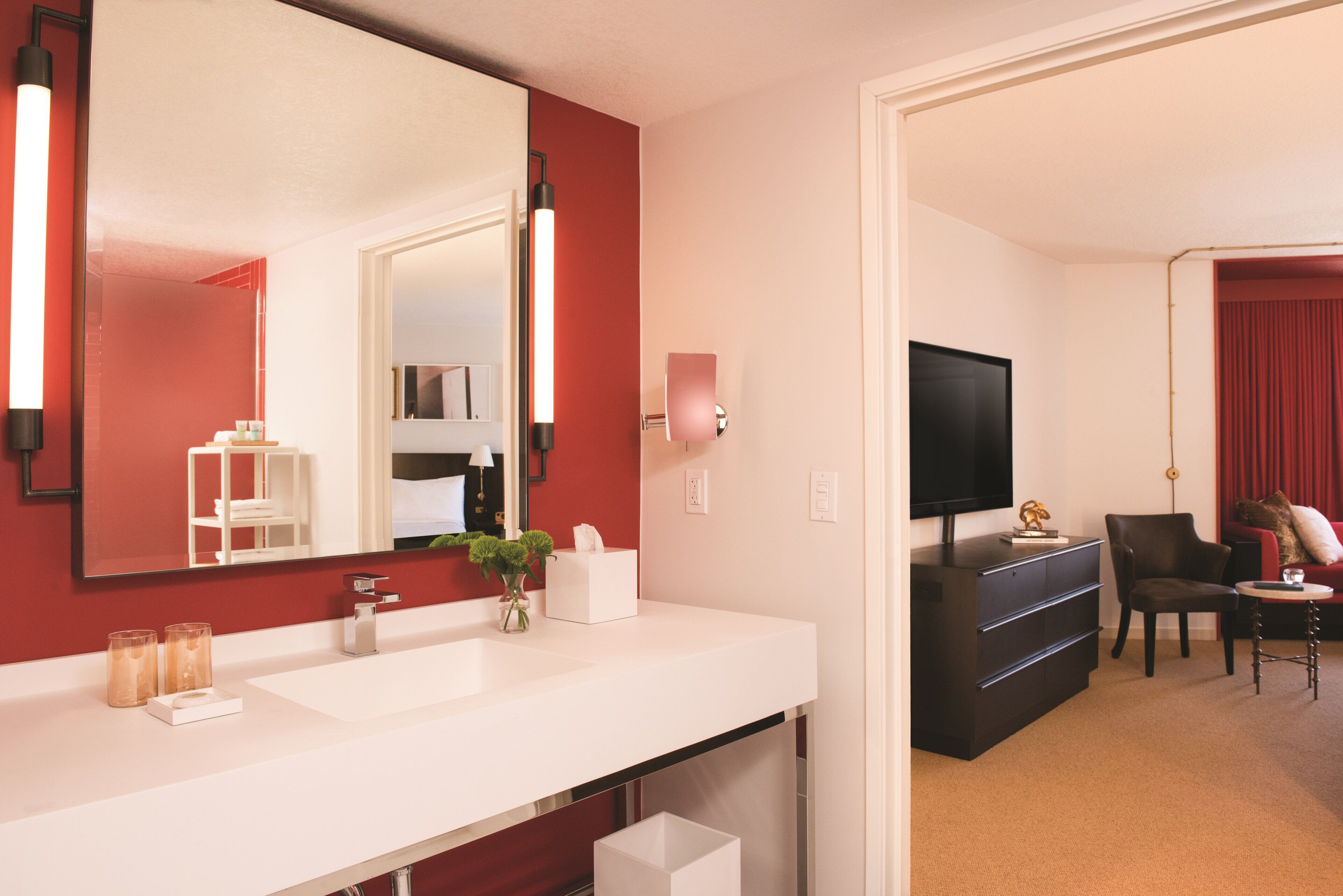 Camera d'albergo moderna con una toeletta bianca, uno specchio grande, una TV a schermo piatto, un comò scuro e un'area salotto con un pannello a tenda rosso.
