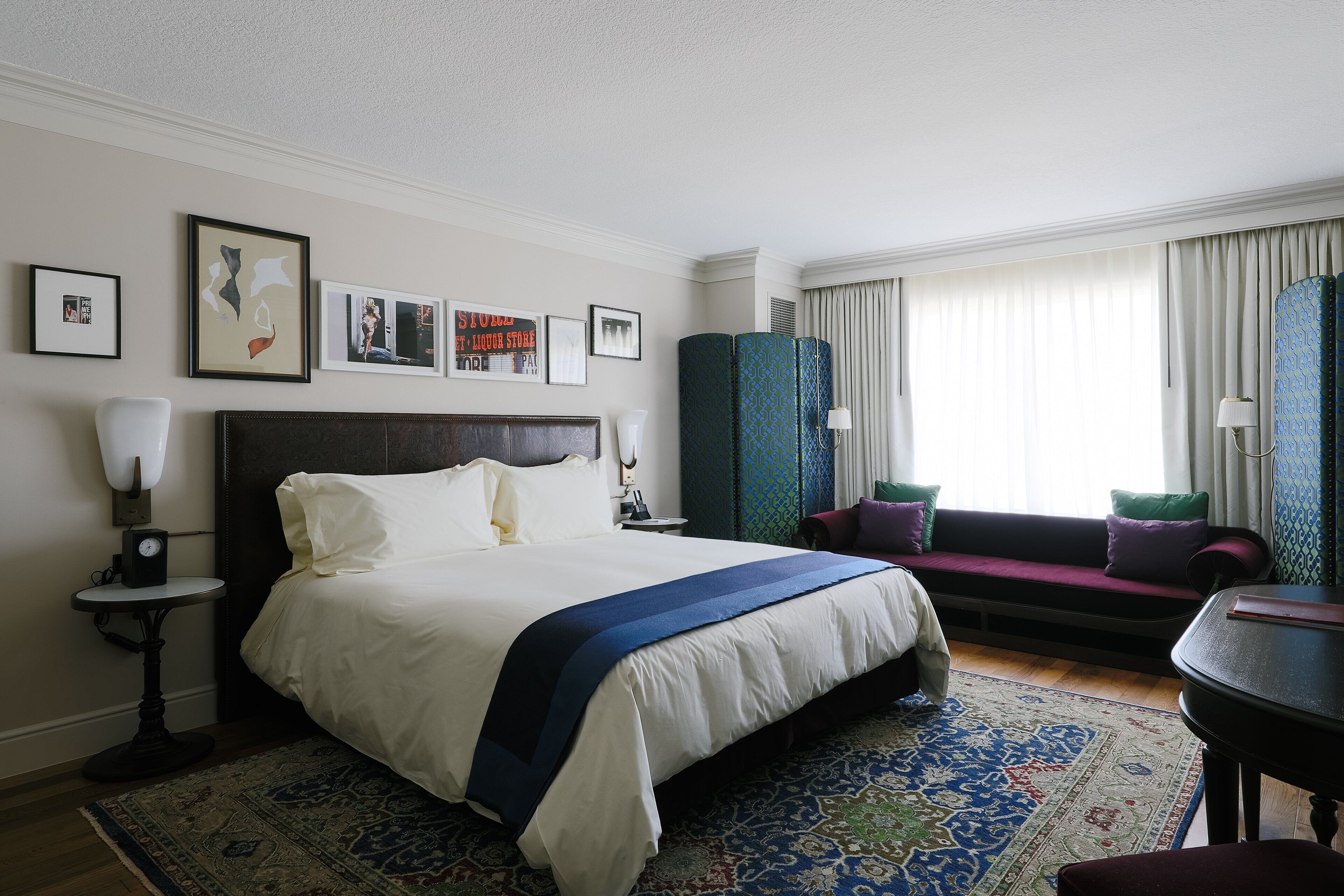 Een hotelkamer met een groot bed, een nachtkastje met lamp, een klein rond tafeltje, een bank met kussens en een groot raam met gordijnen.