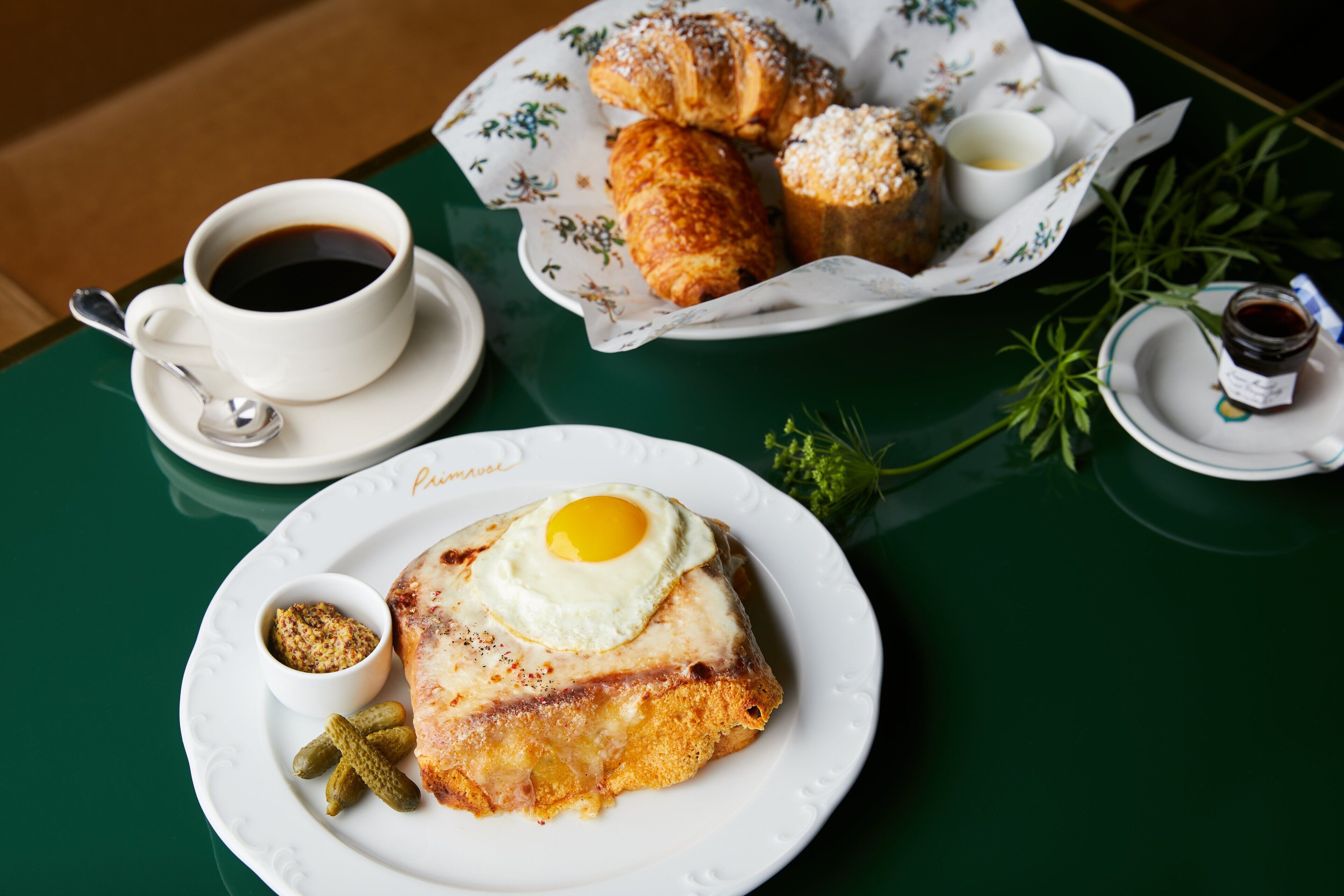 A breakfast setting with a croissant, a cup of coffee, and a plate with a fried egg and pickles.