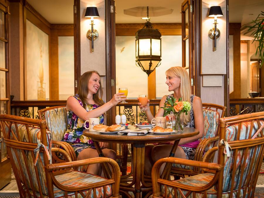 Duas pessoas sentadas em uma mesa redonda com uma garrafa de champanhe, um copo de suco de laranja e um prato de comida. Elas estão em um ambiente com cadeiras de vime e um lustre.