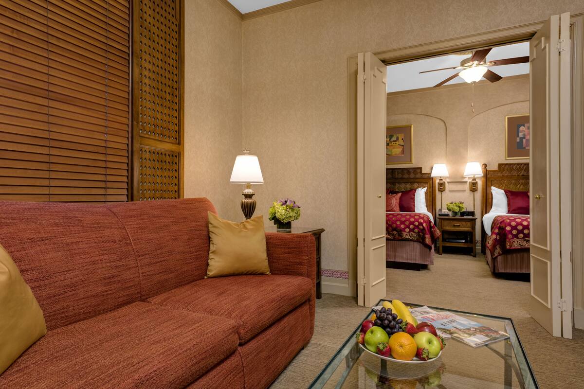 Een hotelkamer met een rode bank, een glazen bijzettafeltje met fruit, een lamp en een plafondventilator.