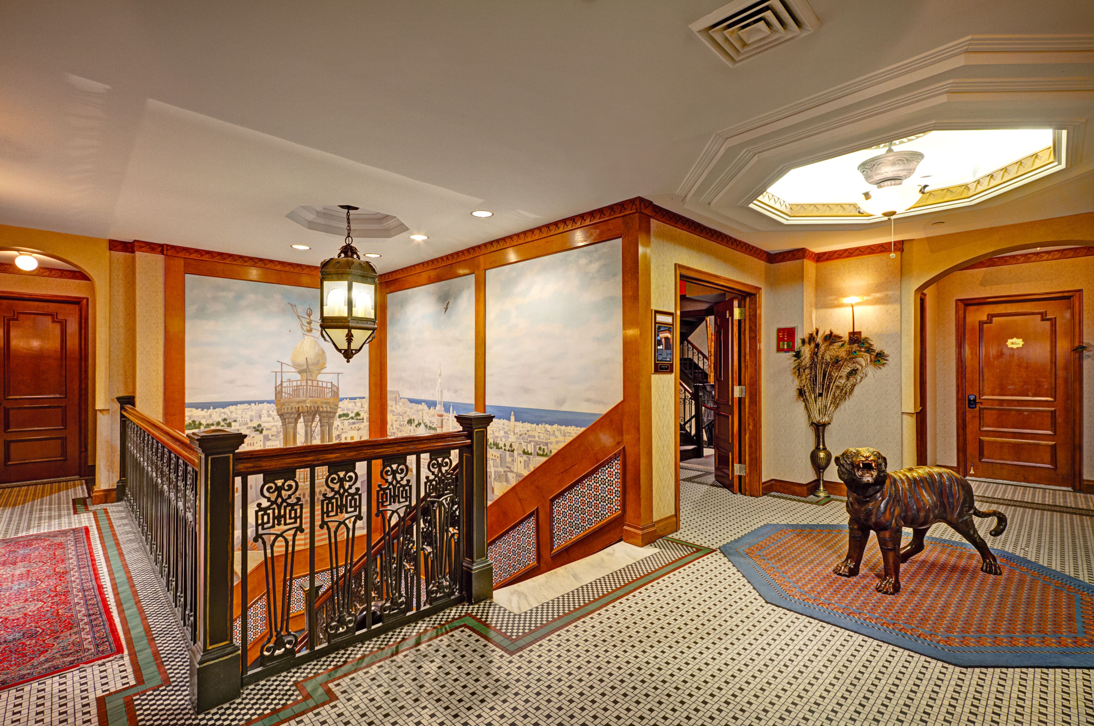 Un vaste hall d’entrée avec un escalier, une fresque représentant un paysage urbain, une statue de chien décorative et un tapis à motifs.