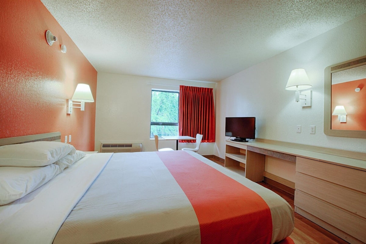 A hotel room with a bed, a desk, a television, and a window with red curtains.