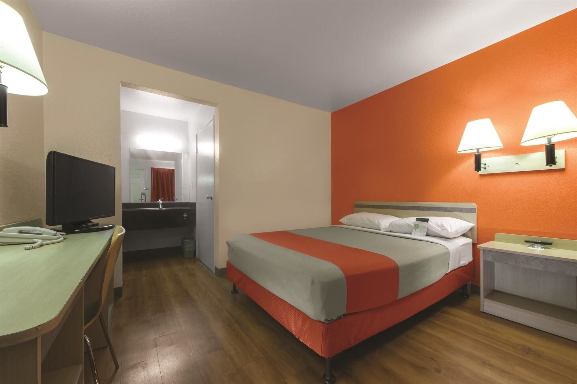 A hotel room with a bed, a desk, a television, and a bathroom visible through an open door.
