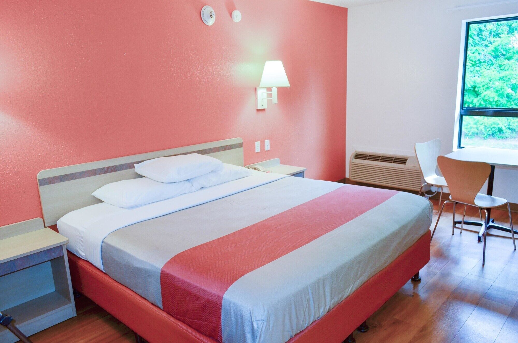 A hotel room with a bed, bedside tables, a desk, a chair, a lamp, and a window with a view of greenery.