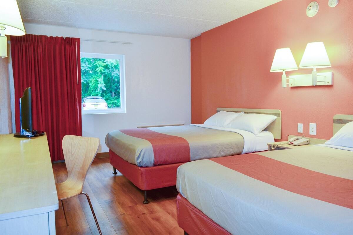 A hotel room with two beds, a desk, a chair, a window with a view of greenery, and a wall clock.