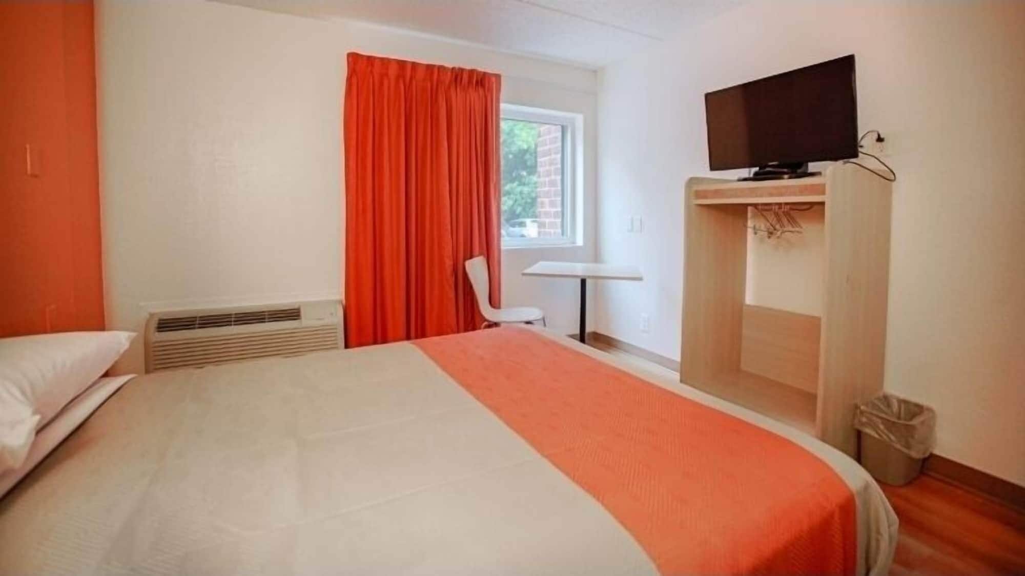A hotel room with a bed, a TV on a stand, a window with curtains, a chair, and a small table.