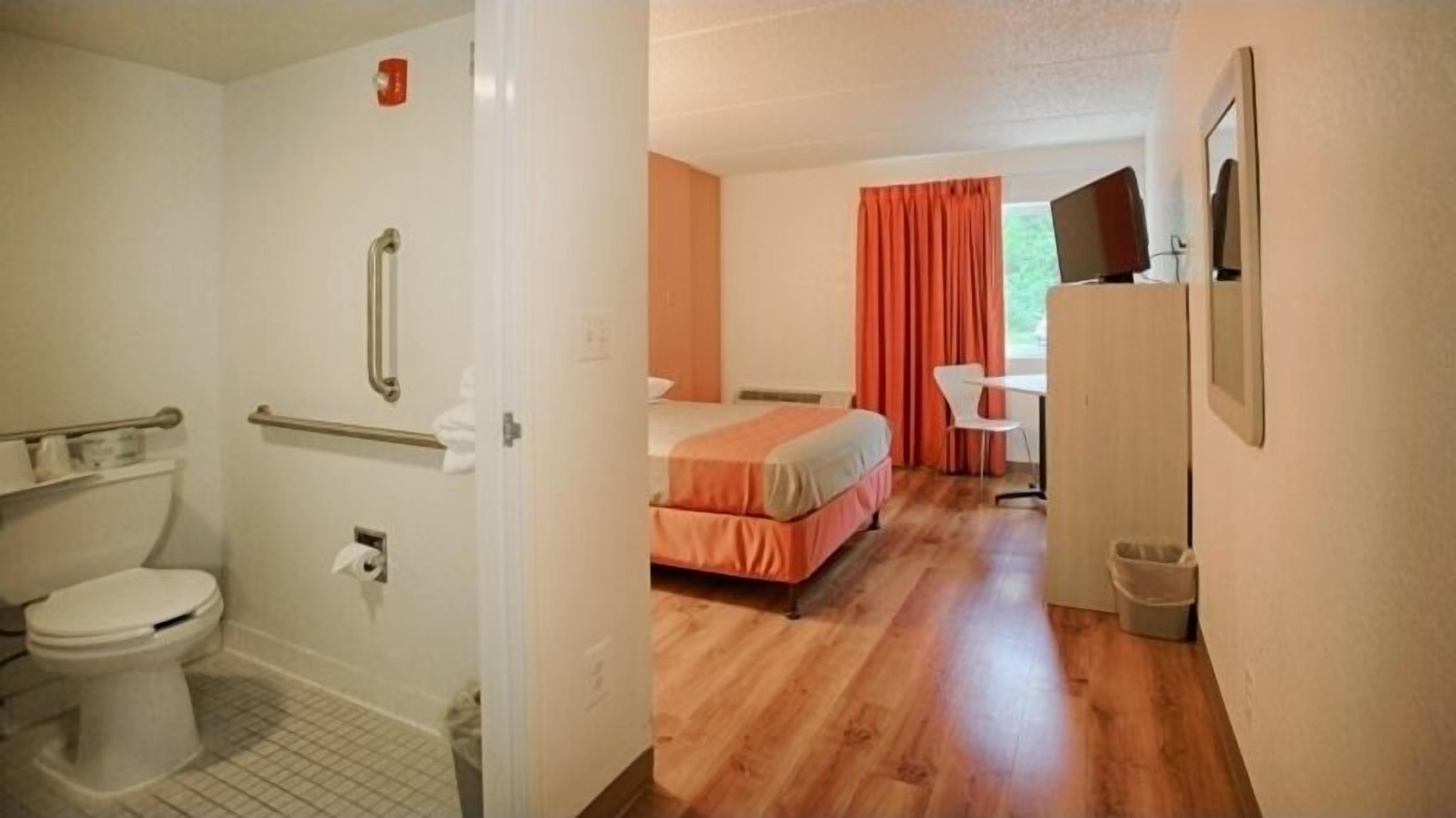 Ein Badezimmer mit Toilette, Haltegriffen und Dusche. Im angrenzenden Raum befindet sich ein Bett mit orangefarbener Bettdecke und ein Fernseher auf einem Schrank.