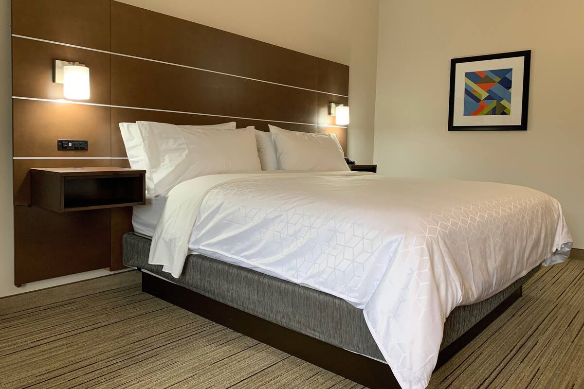 Une chambre d’hôtel dotée d’un grand lit, d’une tête de lit avec éclairage intégré, d’une table de chevet et d’une œuvre d’art abstraite encadrée accrochée au mur.