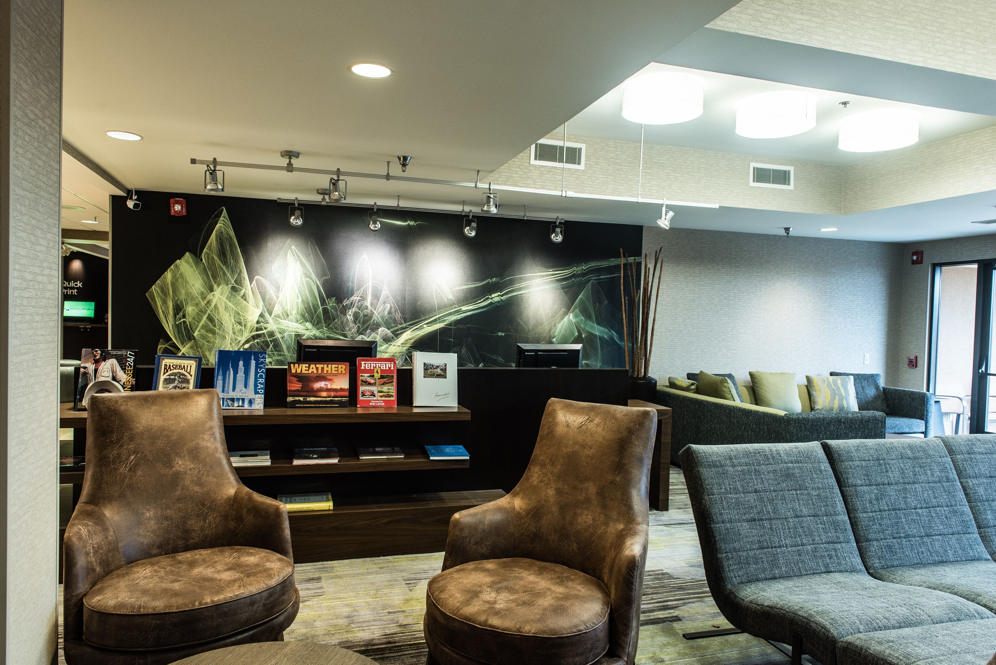 A lobby with two brown leather chairs, a grey upholstered sofa, and a bookshelf with books and decorative items.