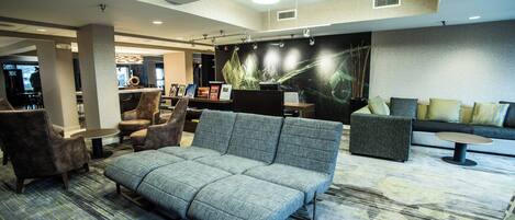 En moderne lobby med en grå sofa, lænestole og et lille bord. Der er en reception med en computer og en stor plante.