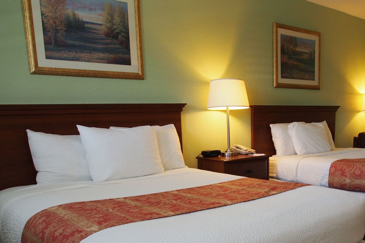 A hotel room with two beds, a nightstand with a lamp, and two framed landscape paintings on the wall.