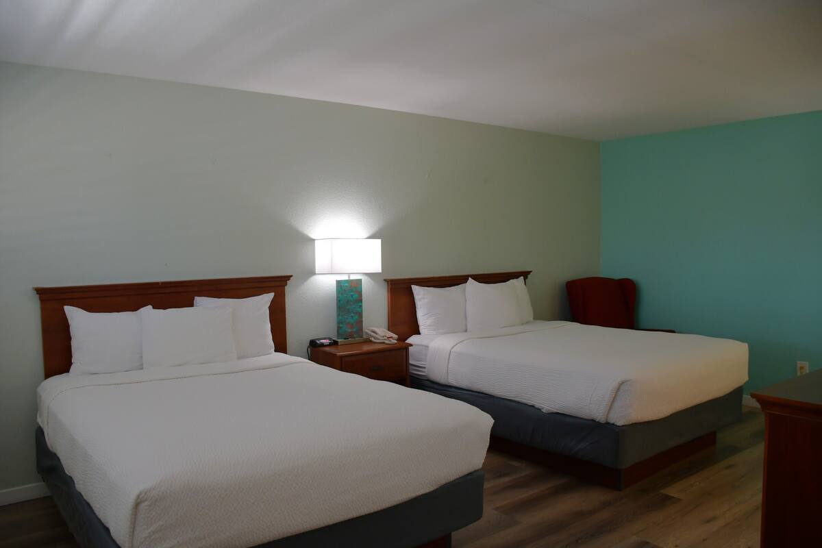 A hotel room with two beds, wooden headboards, a nightstand with a lamp, and a teal accent wall.