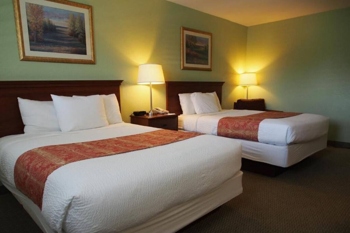 Two queen beds in a hotel room with headboards, bedside lamps, and framed artwork on the walls.