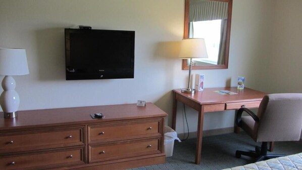 A hotel room with a wooden desk, a television mounted on the wall, a lamp, a chair, and a trash bin.