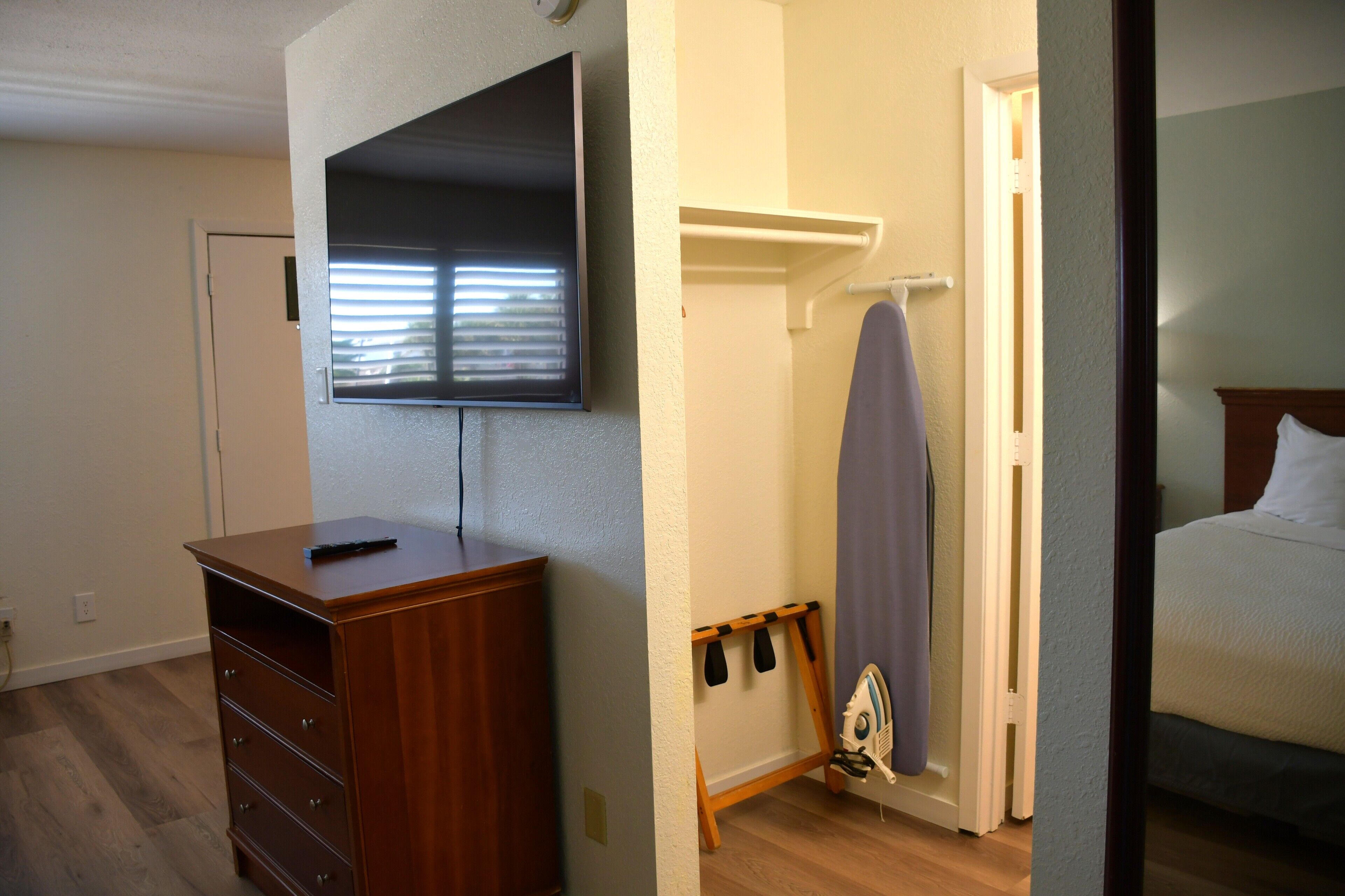 A bedroom with a wooden dresser, a flat-screen TV mounted on the wall, a shower with a towel rack, and a bed with white bedding.