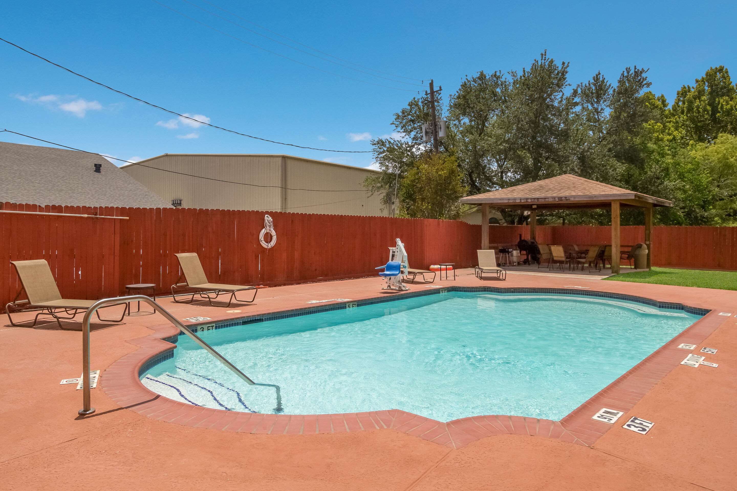 A rectangular swimming pool with a metal ladder, surrounded by a red fence. There are lounge chairs and a covered seating area in the background.