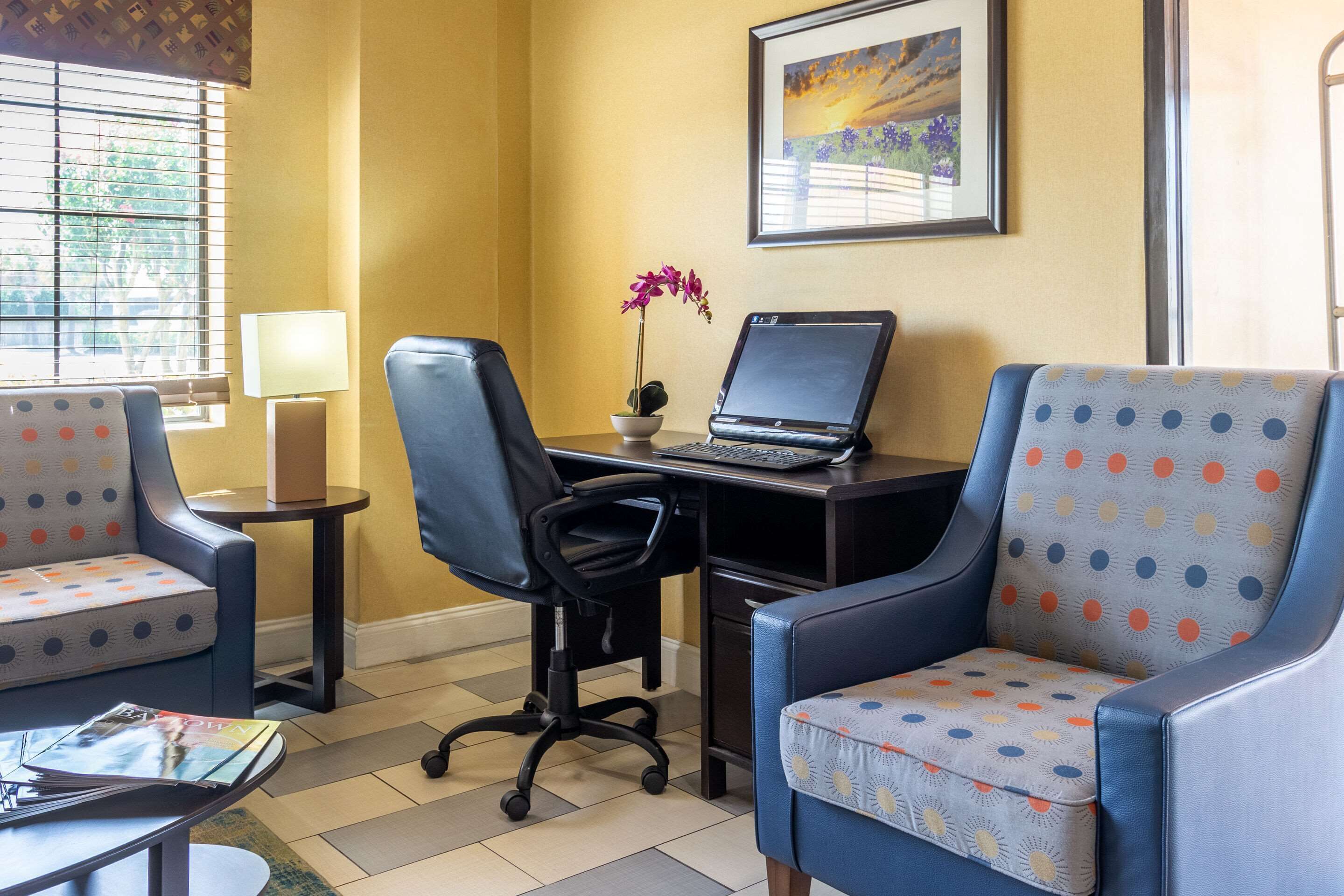 A hotel room with two blue armchairs, a desk with a laptop, a small table with a lamp, and a window with blinds.