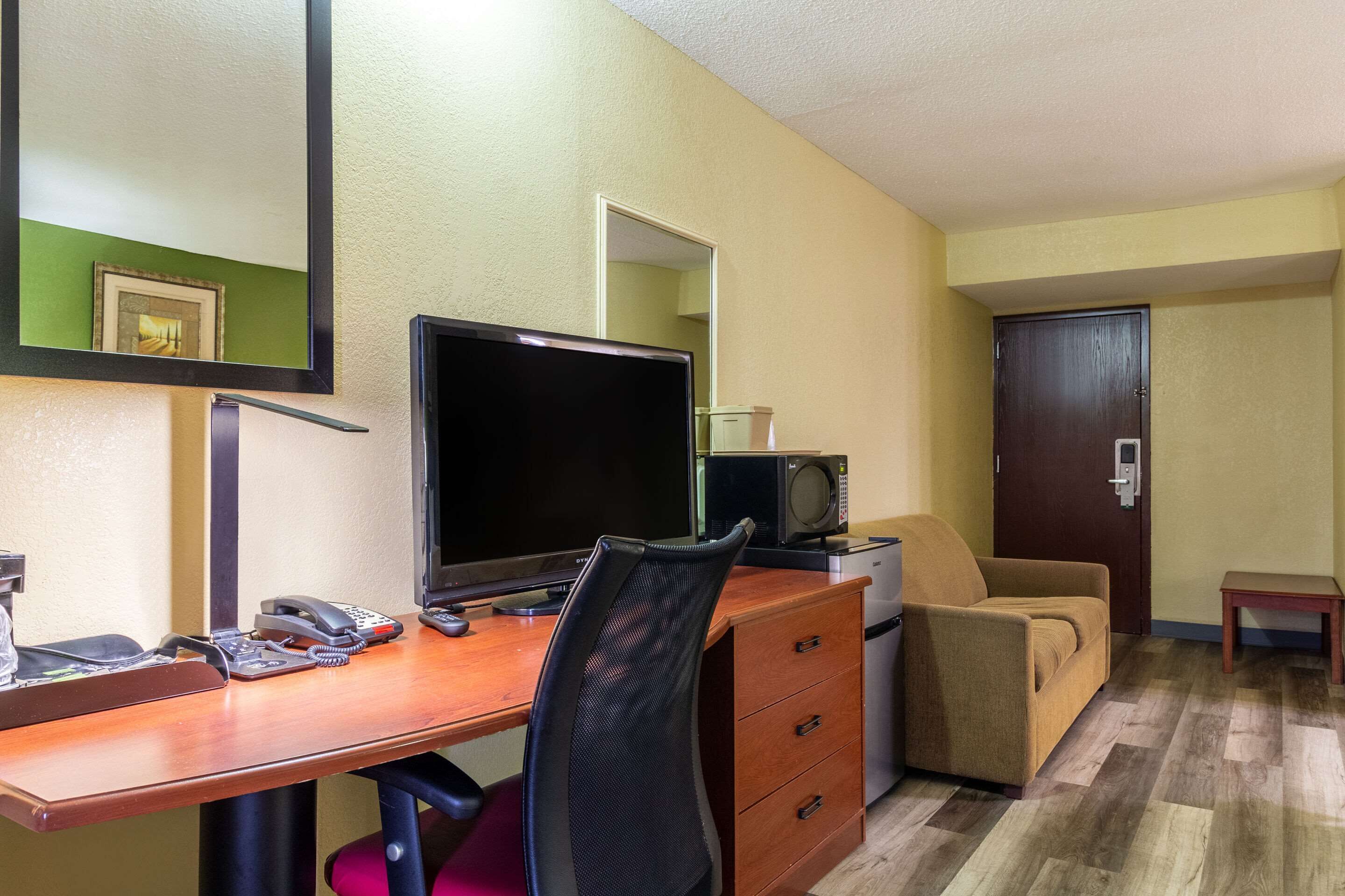A hotel room with a wooden desk, a computer, a telephone, a microwave, a chair, a sofa, and a door.