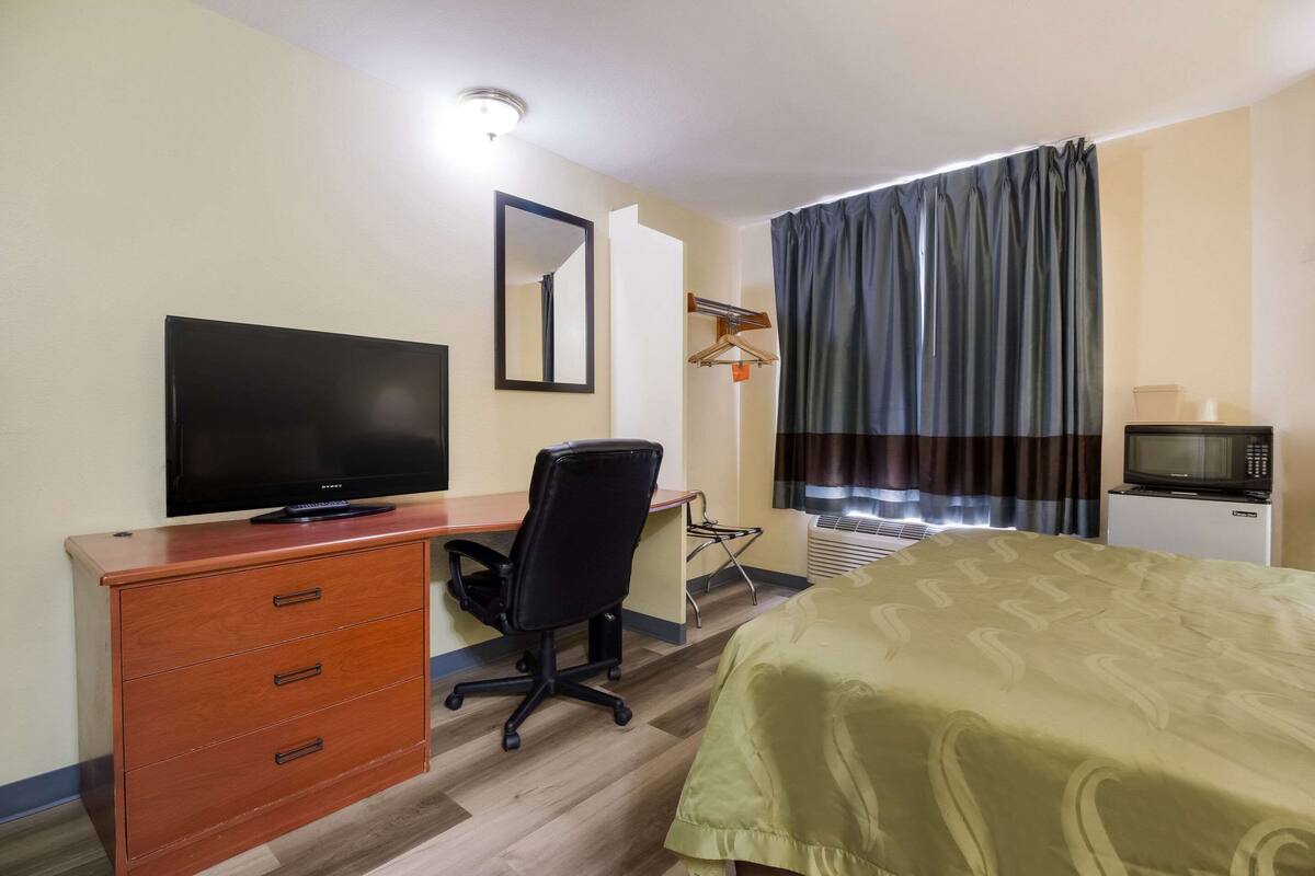 A hotel room with a bed, a desk with a television, a dresser, a chair, and a small kitchenette.