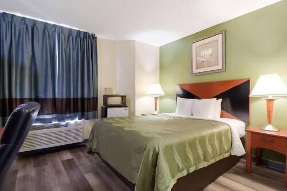 A hotel room with a bed, two bedside lamps, a desk with a chair, a television, and a framed picture on the wall.