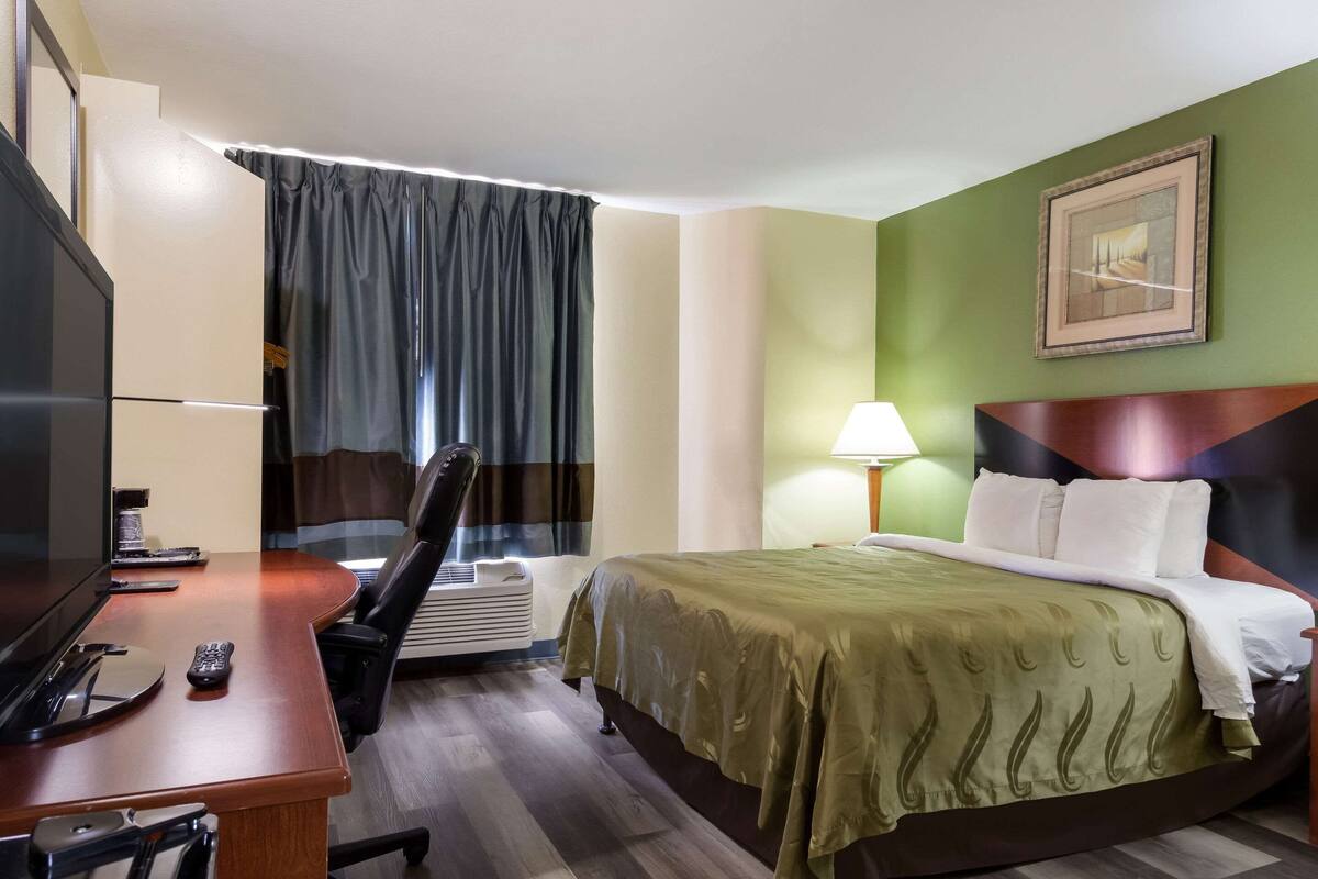 A hotel room with a bed, a desk with a TV, a chair, a lamp, and a painting on the wall.