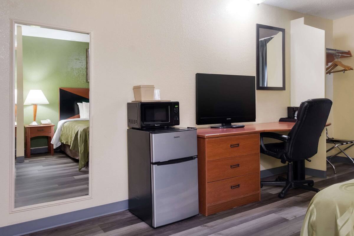 A hotel room with a bed, a desk with a computer, a microwave, a refrigerator, a dresser, and a mirror.