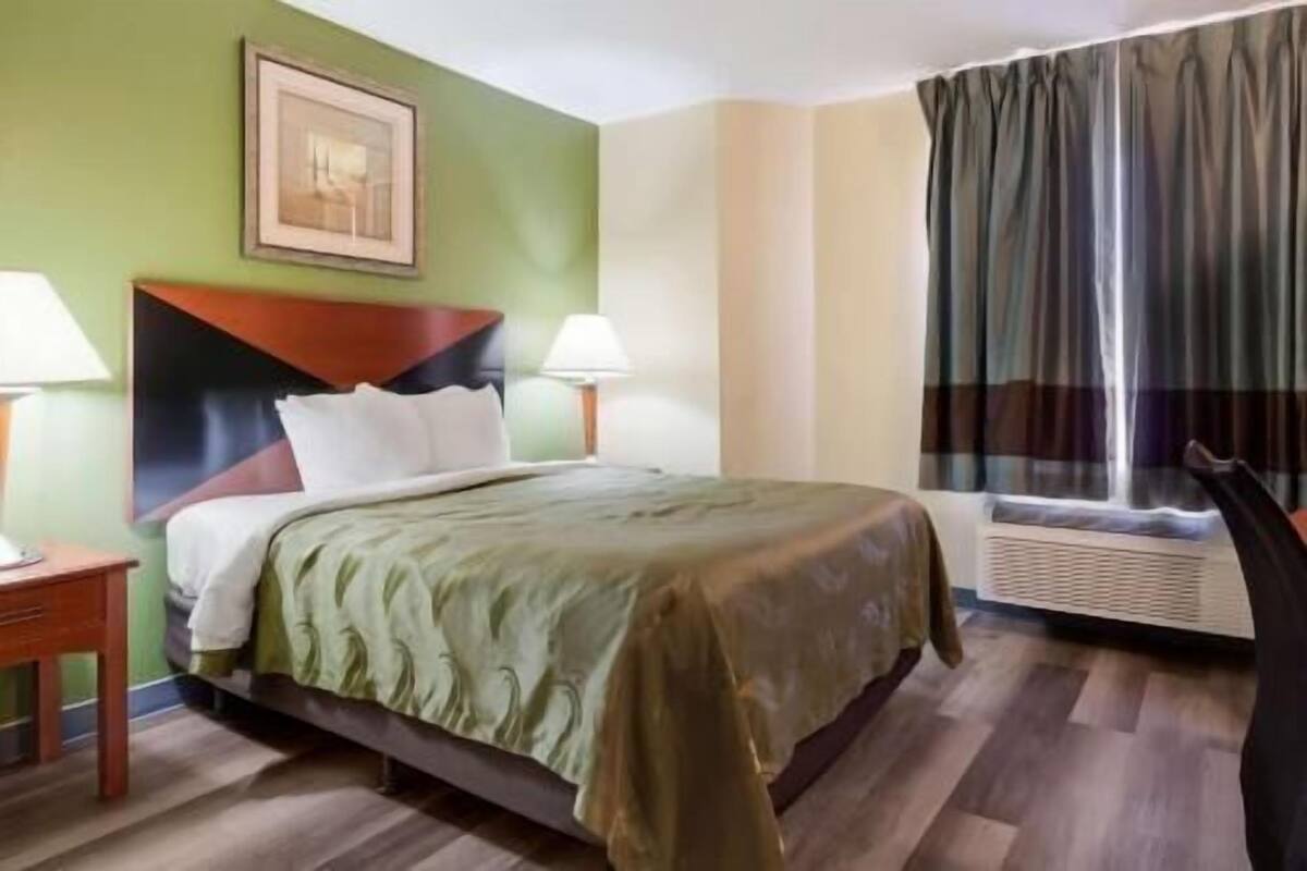 A hotel room with a bed, two bedside lamps, a window with curtains, a radiator, and a framed picture on the wall.