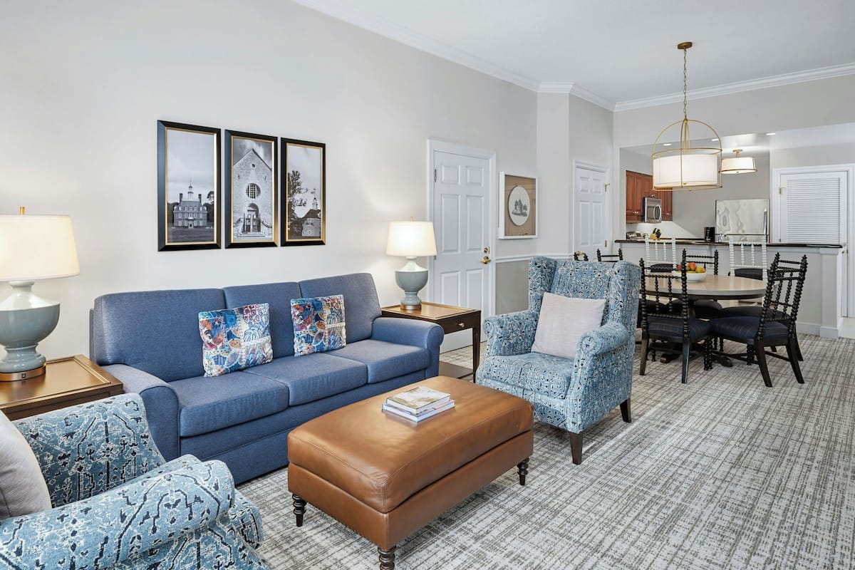 A living room with a blue sofa, patterned armchairs, a brown ottoman, and a dining area with a round table and chairs.