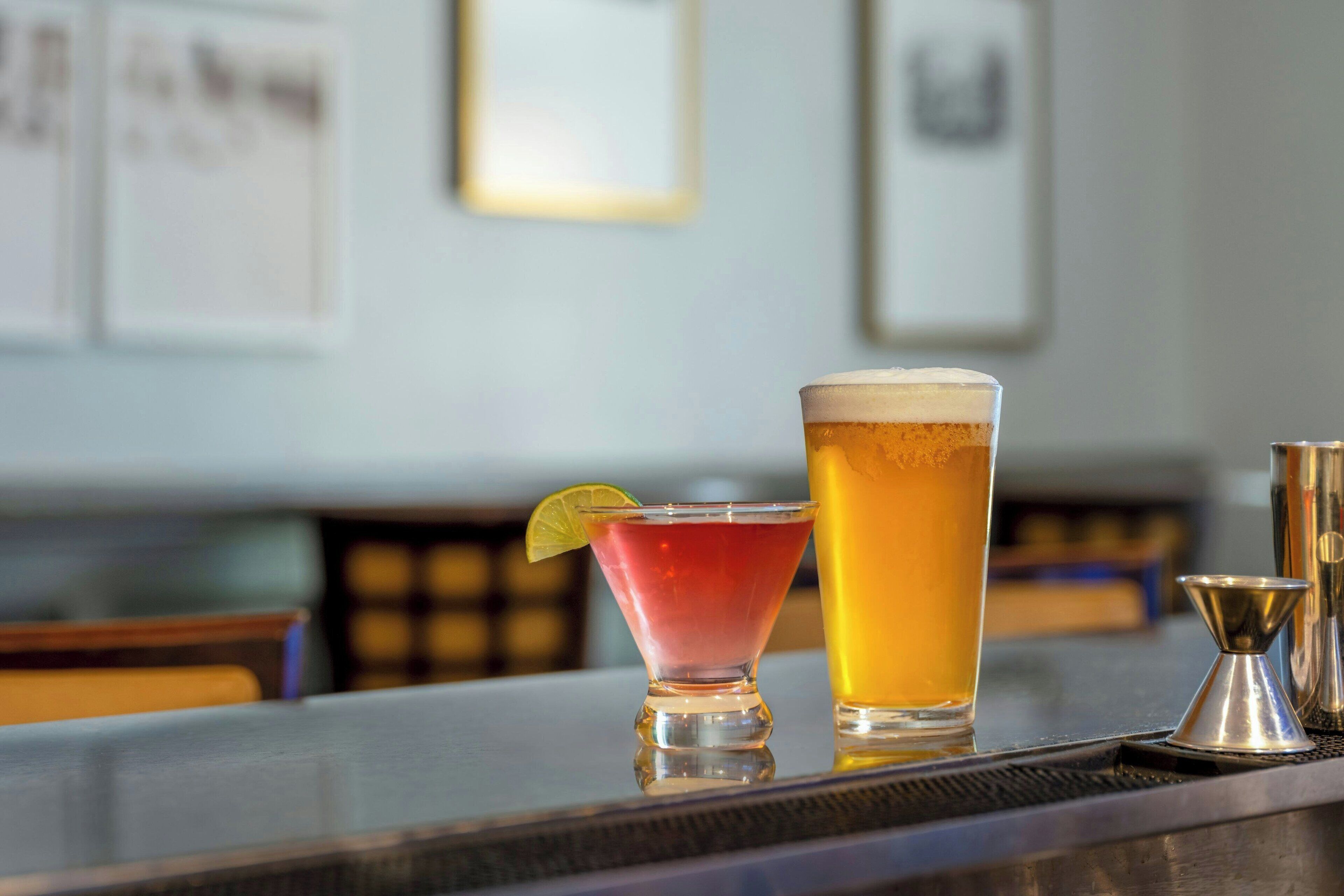 Deux verres de bière sur le comptoir d’un bar, avec une tranche de citron vert, un shaker en métal et un panneau de menu en arrière-plan.